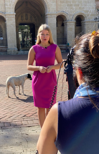 One person stands in front of building, talking to camera, while another person records.