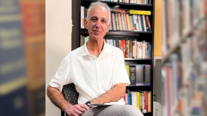 Adam Braver sitting in a chair in front of a bookcase full of books. 