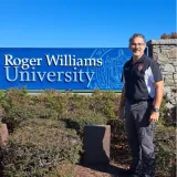 Alumni John Scott standing in front of the Roger Williams University sign outside on a day with a blue clear sky.